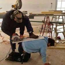 Photo of a man using other man as a table, so he can cut a board with a buzz saw